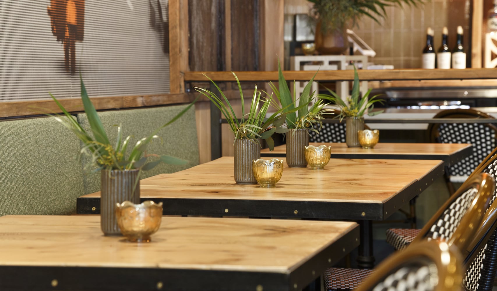 Candles and plants line a row of restaurant tables, wine bar in the background